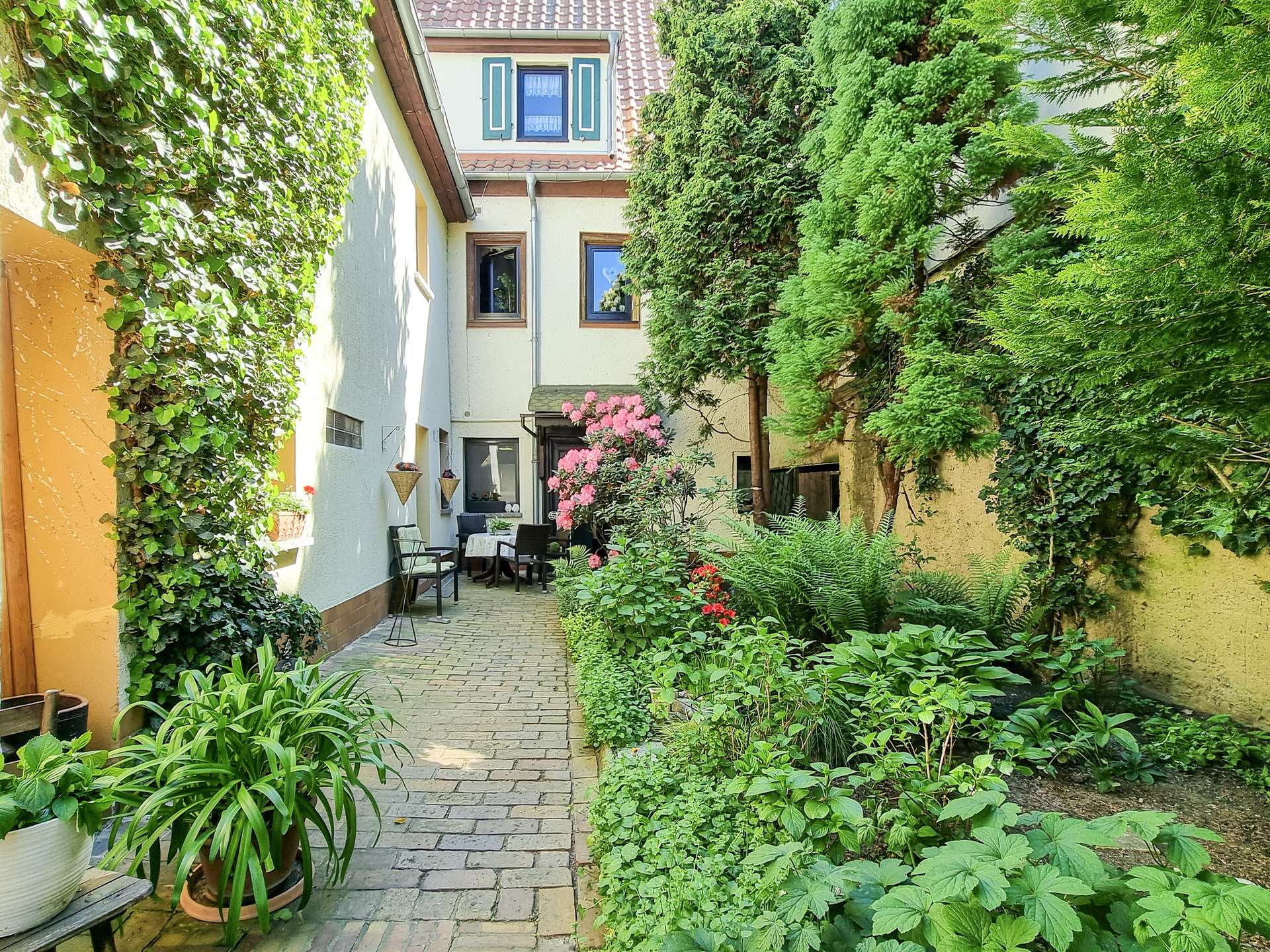 Sunlit narrow courtyard with ivy-covered walls, potted plants, and a small dining table with chairs along a brick path.