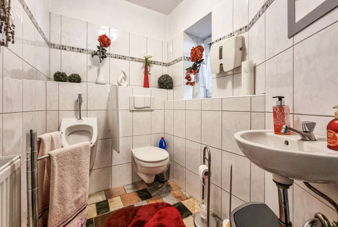 White tiled bathroom with a sink, toilet, and red artificial flowers on a shelf along the wall. A red rug and towels hang nearby.
