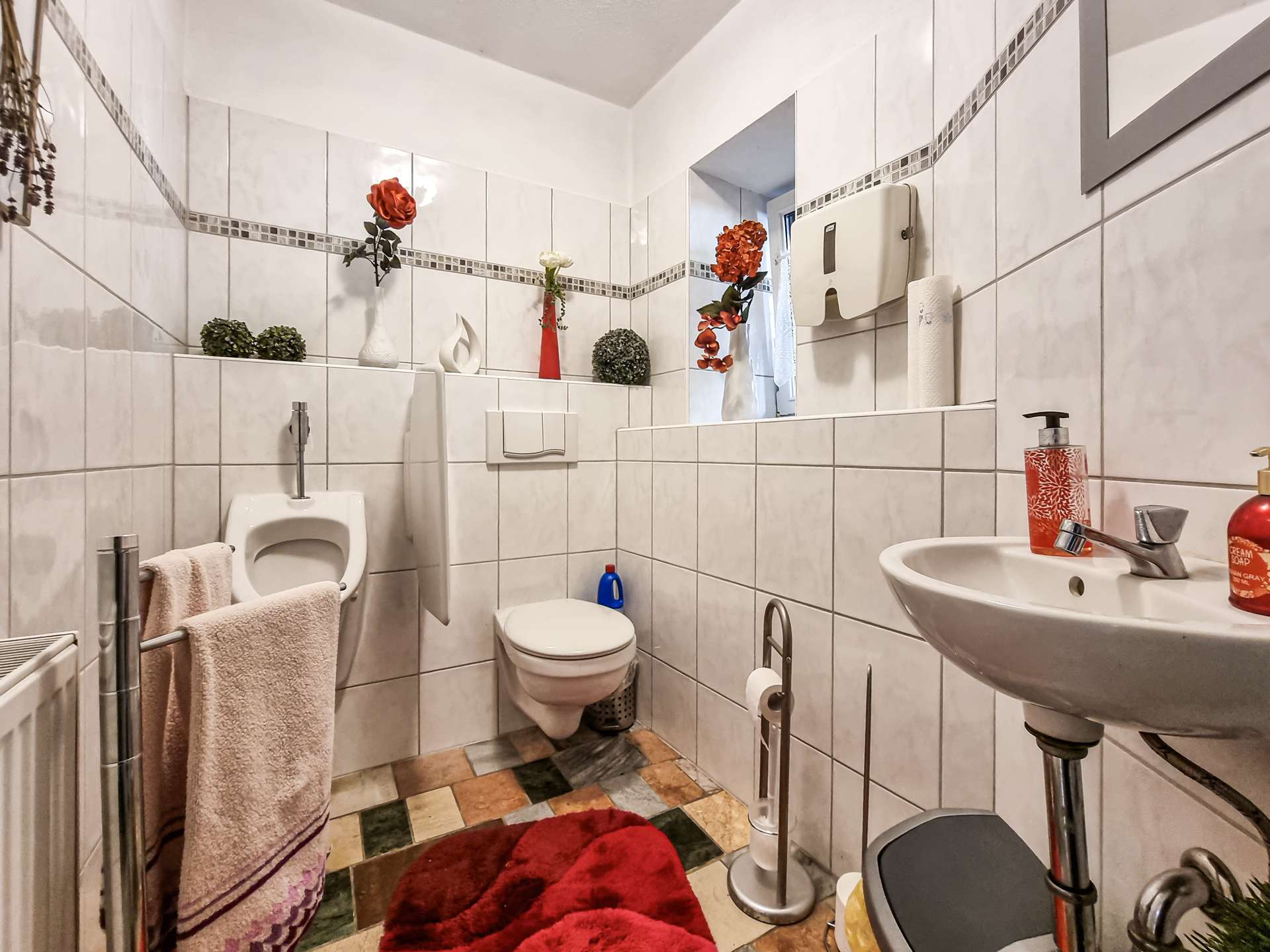 White tiled bathroom with a sink, toilet, and red artificial flowers on a shelf along the wall. A red rug and towels hang nearby.