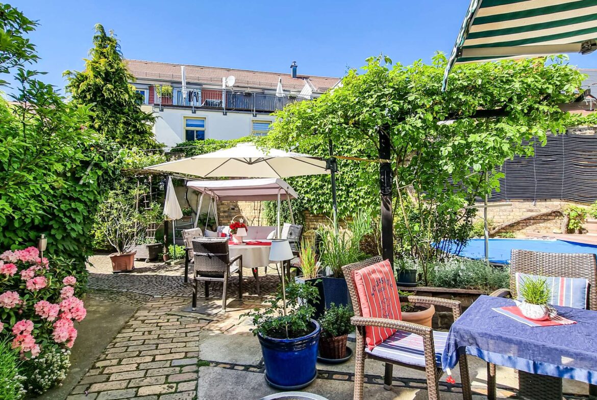 Backyard patio with outdoor dining table, chairs, umbrellas, and lush green plants; residential building with balconies in the background.