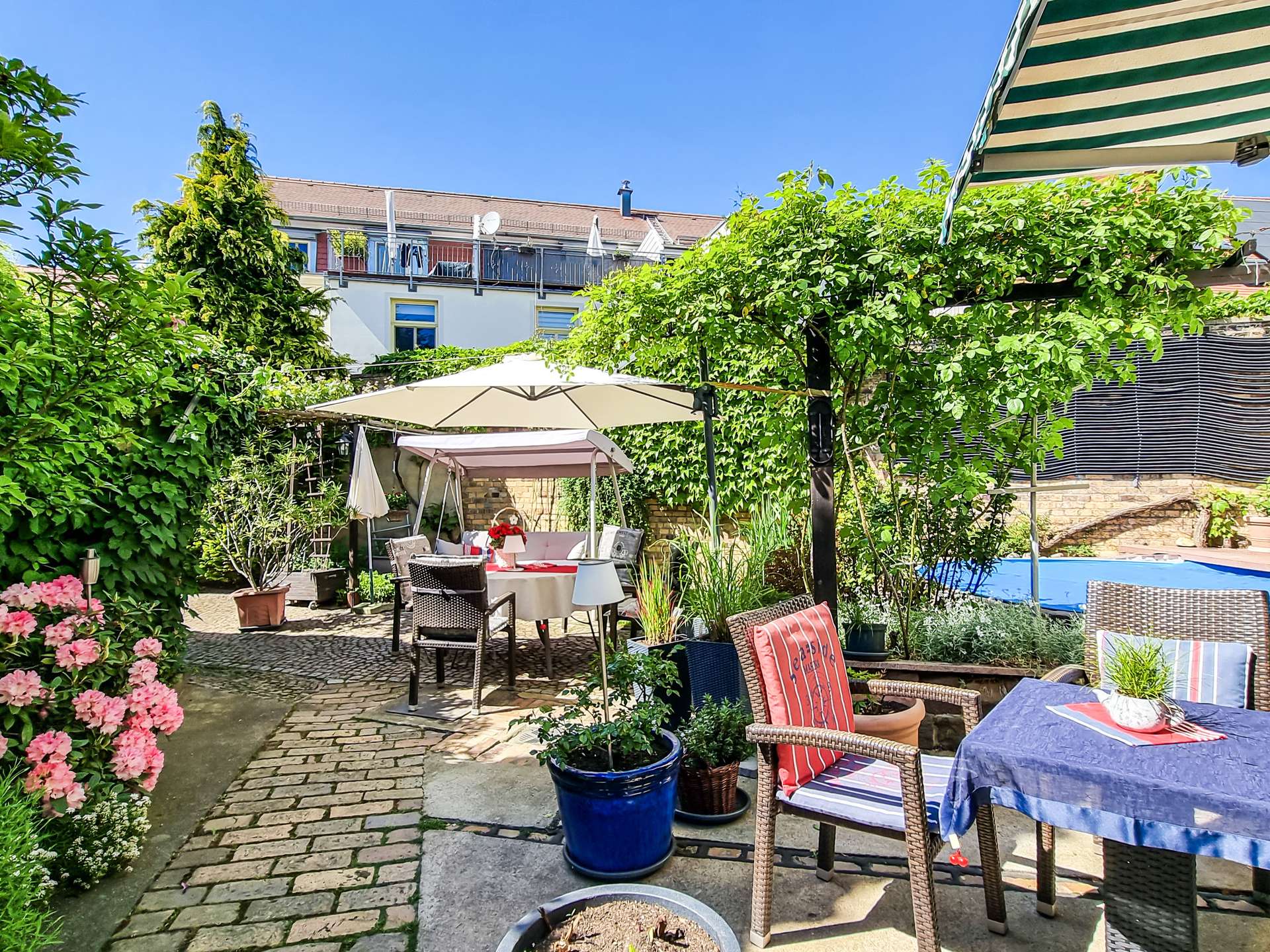 Backyard patio with outdoor dining table, chairs, umbrellas, and lush green plants; residential building with balconies in the background.