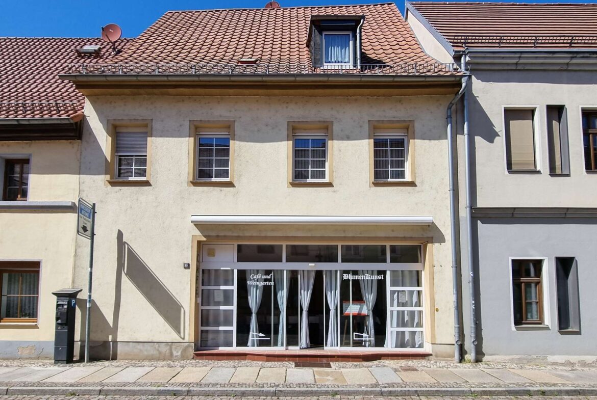 Beige building with a glass storefront, white curtains, and signs BlumenKunst / Café und Weingarten on a cobblestone street.