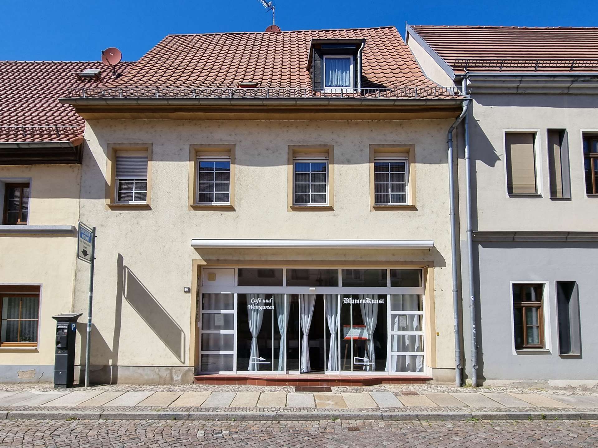 Beige building with a glass storefront, white curtains, and signs BlumenKunst / Café und Weingarten on a cobblestone street.