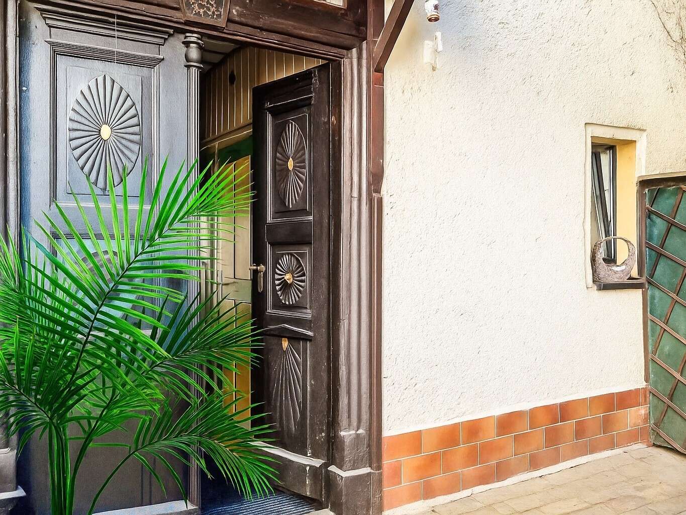 Dark ornate front door with sunburst panels beside a white stucco wall; a large green palm plant dominates the foreground.