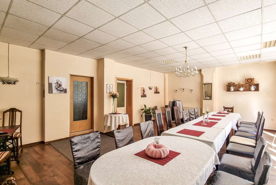 Banquet hall with long white-clothed tables, red placemats, pink napkin centerpieces, gray chairs, and a chandelier in a beige-walled room.