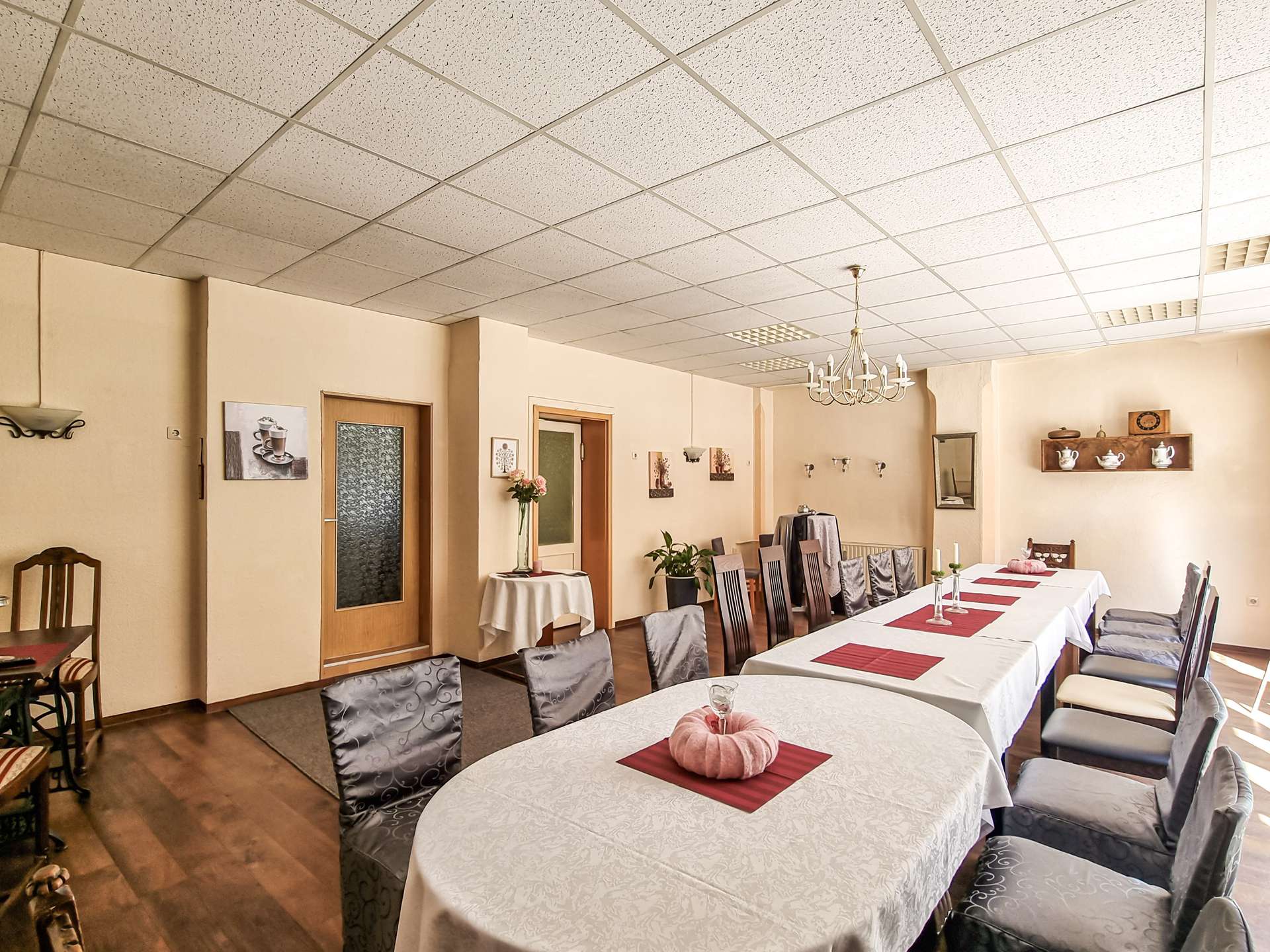 Banquet hall with long white-clothed tables, red placemats, pink napkin centerpieces, gray chairs, and a chandelier in a beige-walled room.