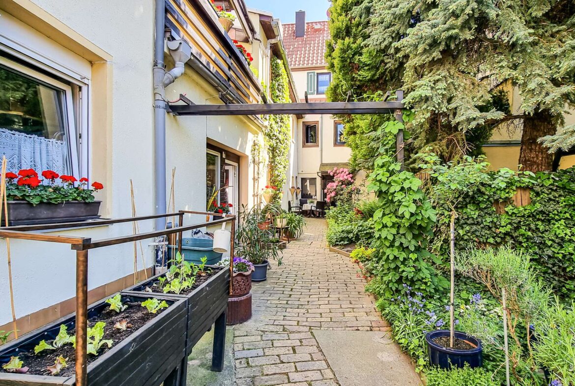 Narrow brick-paved alley between white houses with window boxes and abundant potted plants on both sides.