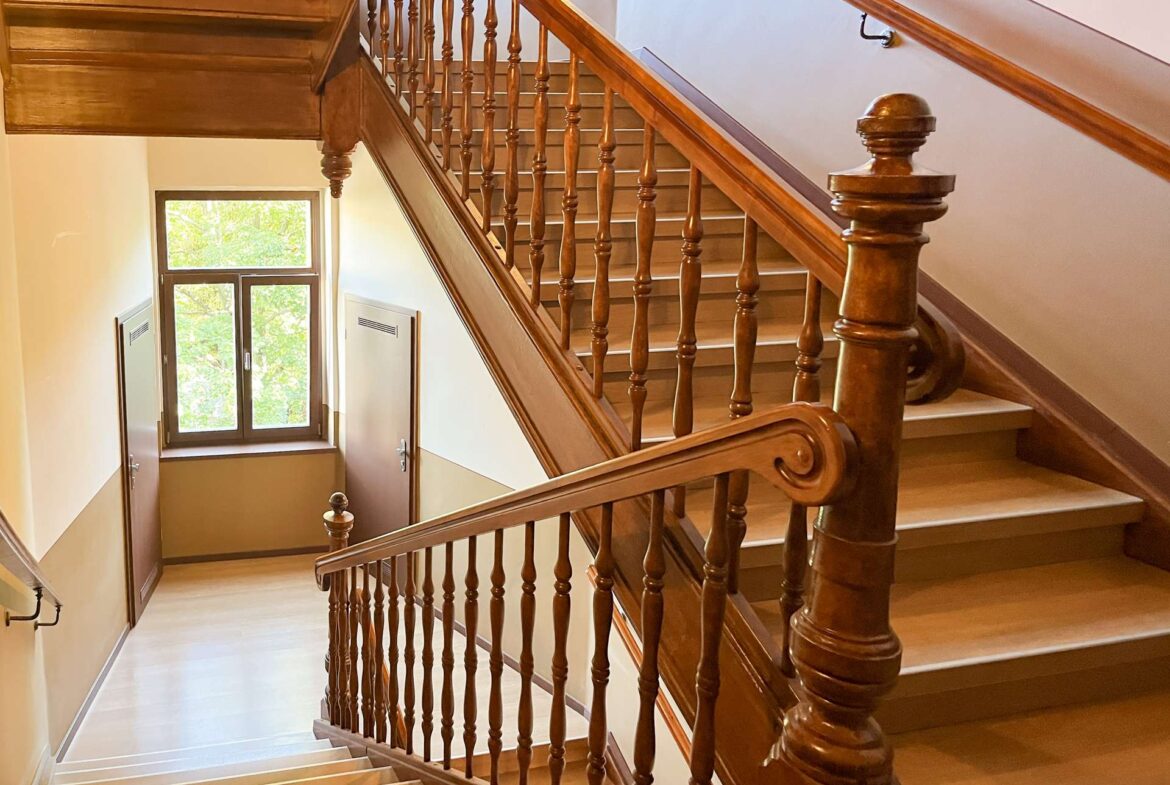 Wooden staircase with ornate banisters inside a building, viewed from mid-staircase looking toward a windowed door area below.