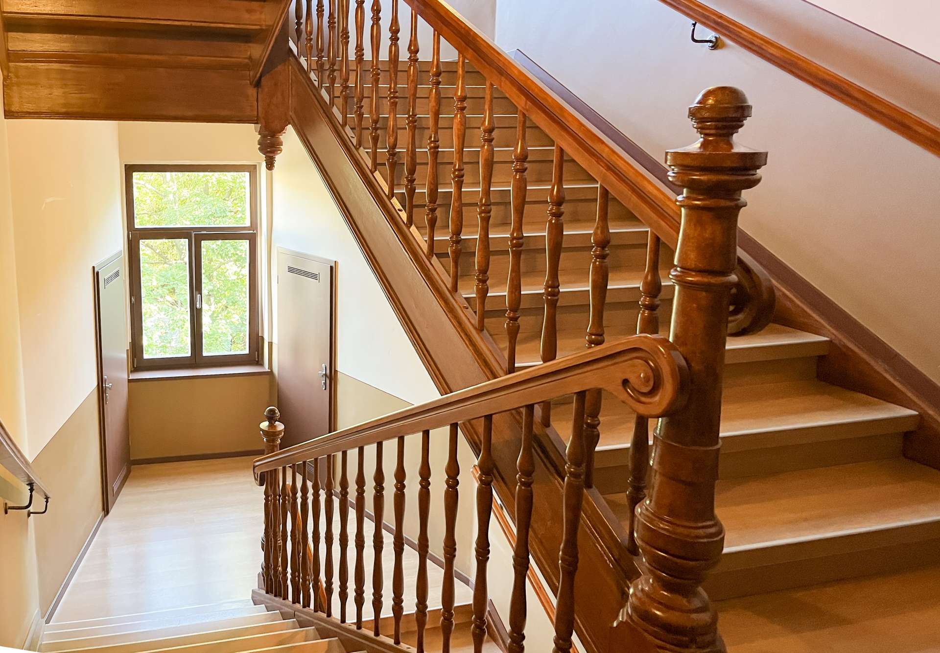 Wooden staircase with ornate banisters inside a building, viewed from mid-staircase looking toward a windowed door area below.