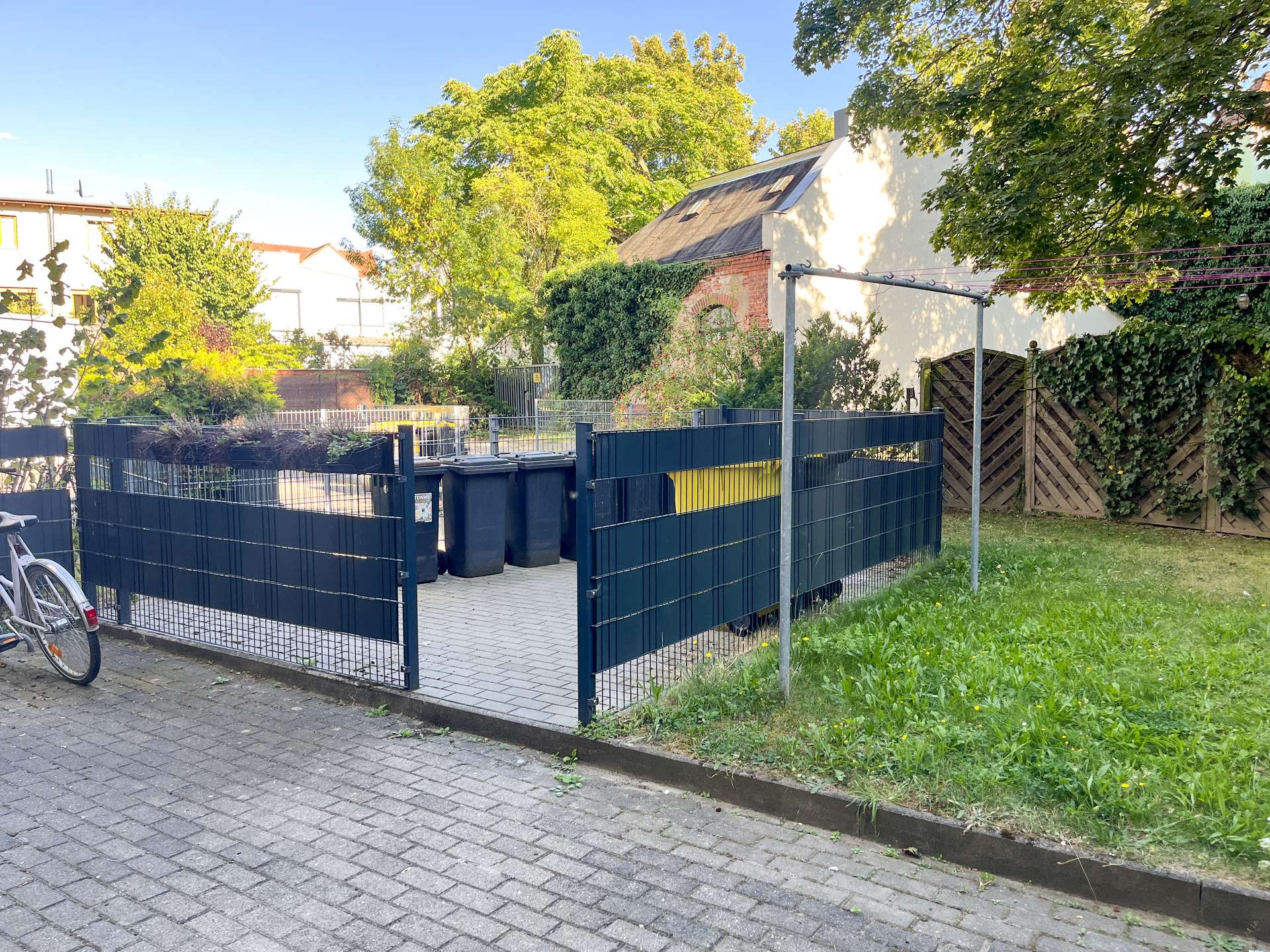 Residential backyard with blue metal fencing, a row of garbage bins, and a bicycle parked at the curb on the left.