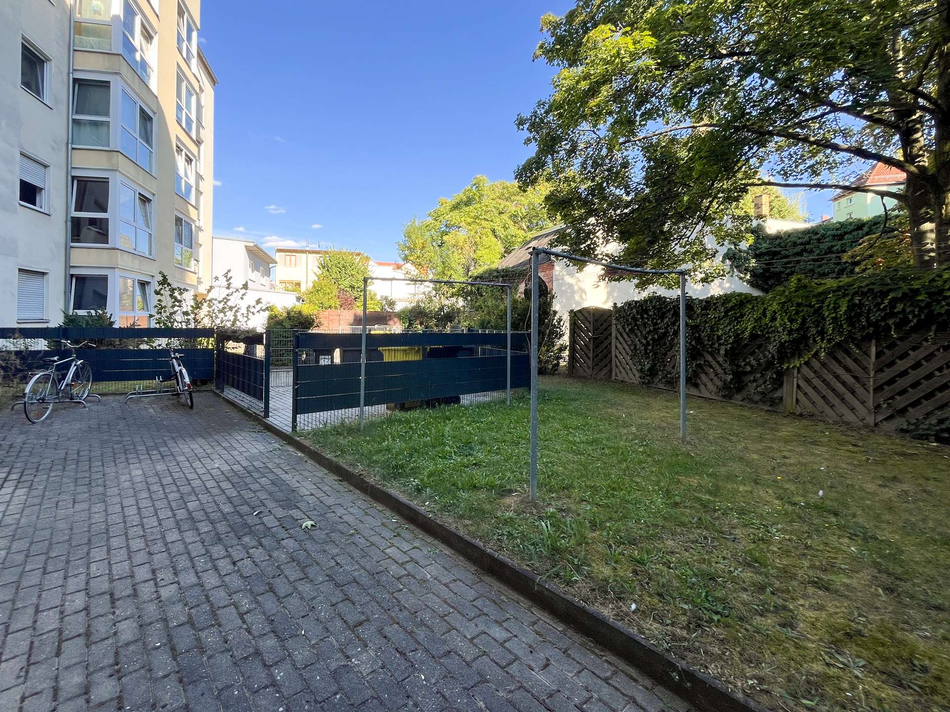 Residential courtyard with a paved brick path, bicycles, and a green fence beside a modern apartment building under a clear blue sky.
