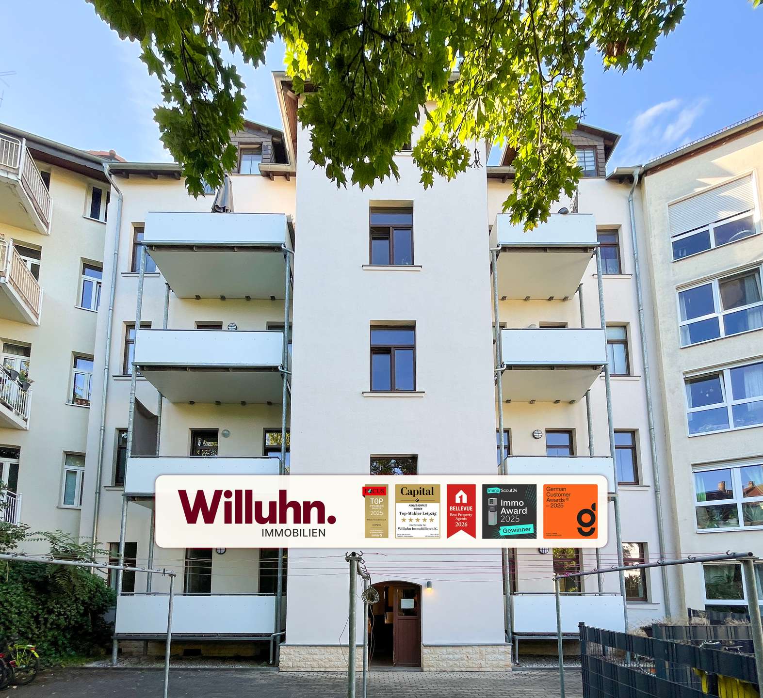 Modern residential building with balconies and a large Willuhn Immobilien sign in front, advertising awards and badges.