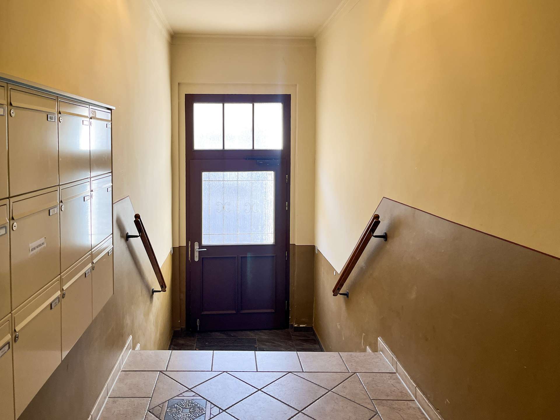 Narrow apartment hallway with beige walls, a row of mailboxes on the left, and a dark front door at the far end.