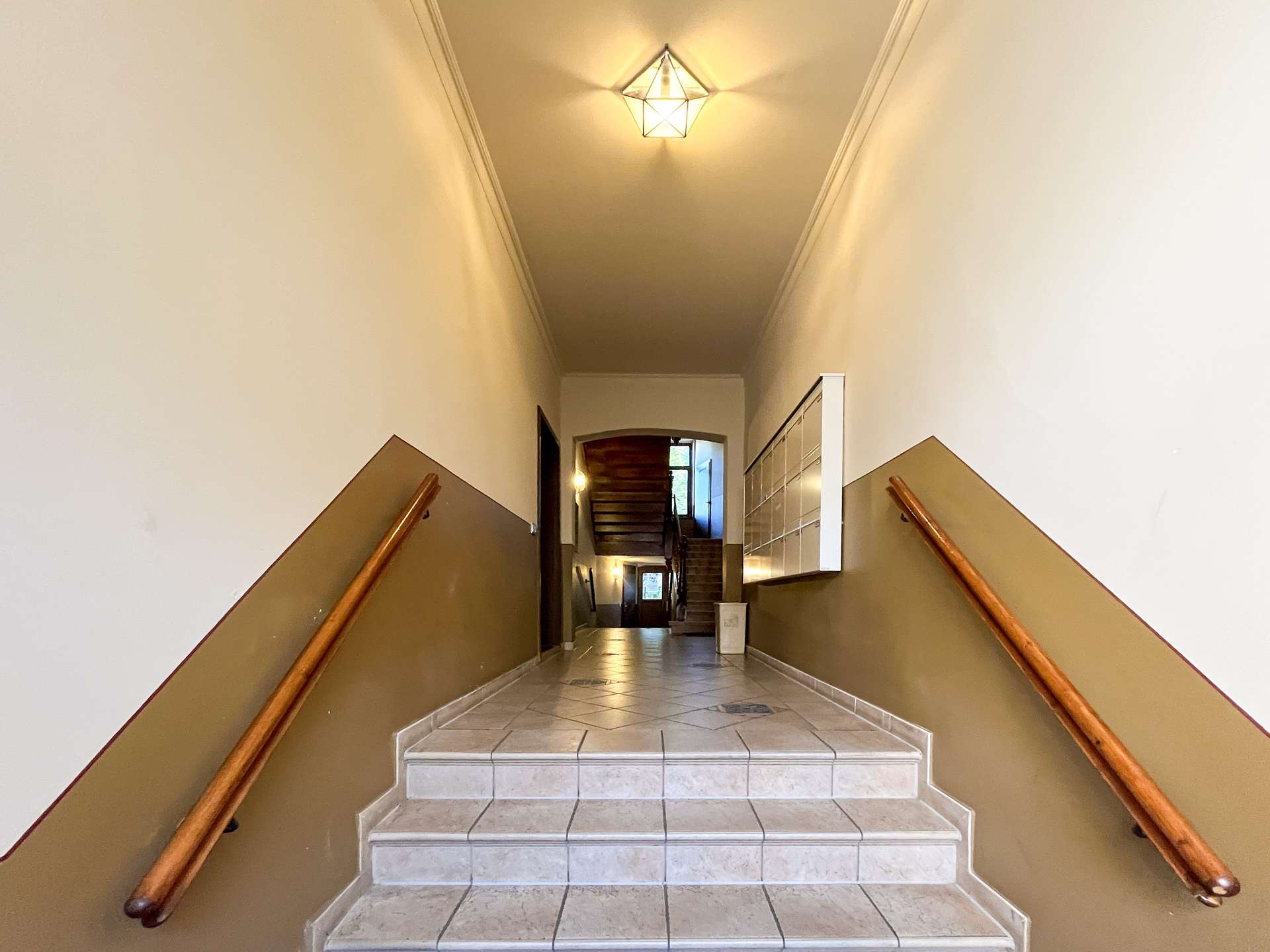 Interior stairwell with tiled steps, brown walls, and wooden handrails on both sides leading to a doorway at the top.