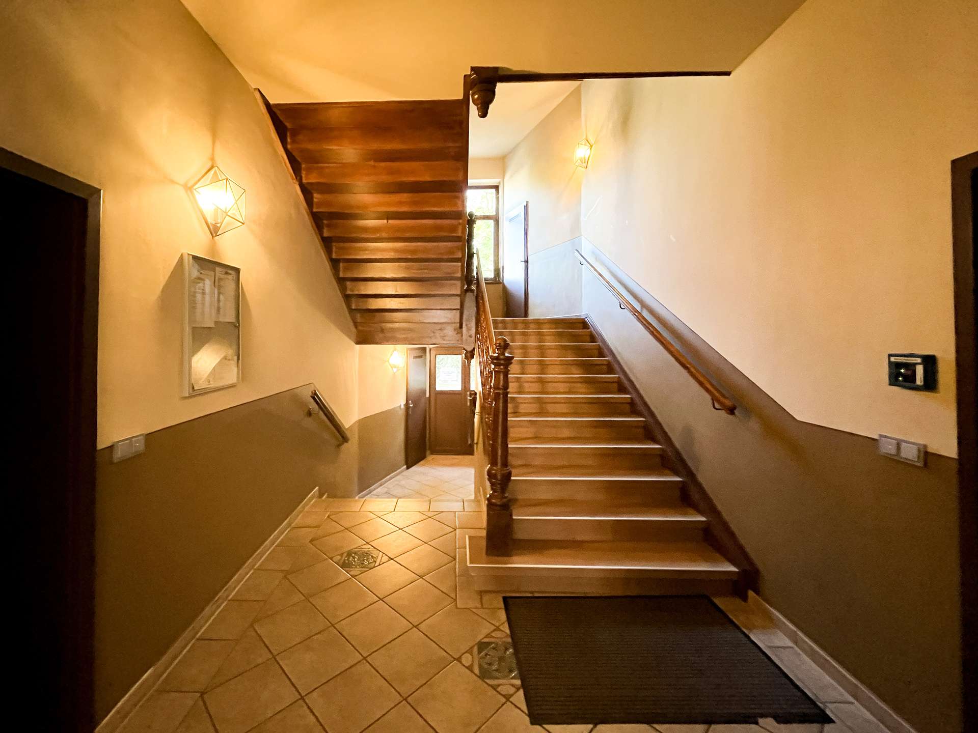 Interior stairwell with wooden stairs and handrail, warm pendant lighting, and tiled floor leading to upper levels.