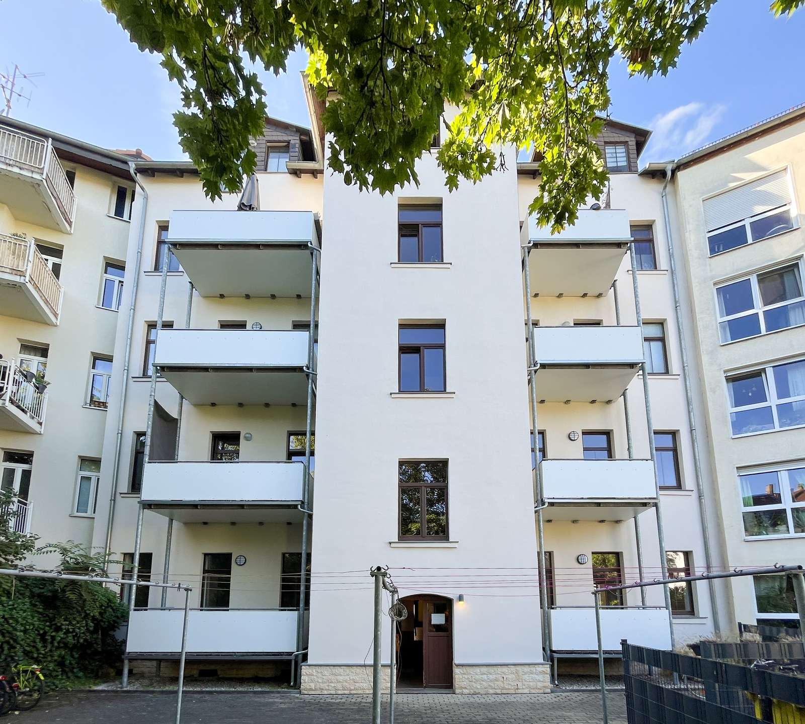 Front view of a white apartment building with multiple balconies and a central tower, shaded by a leafy tree canopy above.