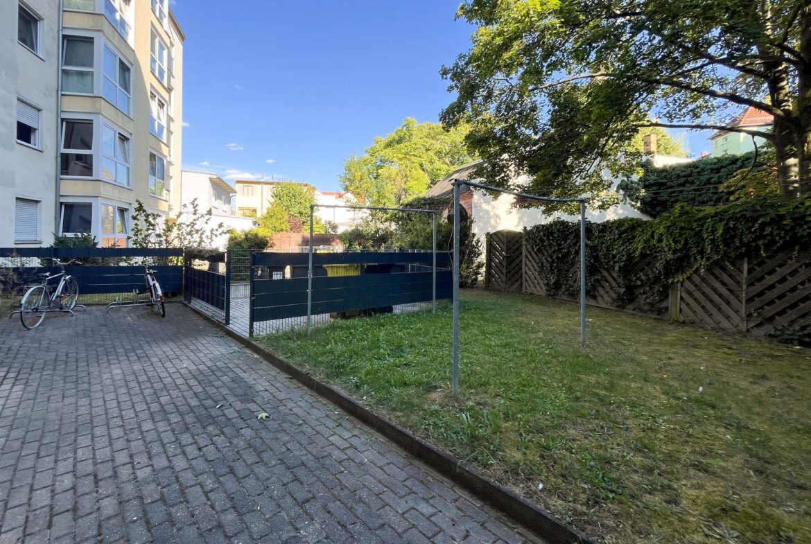 Courtyard of a residential complex: a multi-story beige building to the left, bicycles by a dark fence, and trees over green grass under a blue sky.