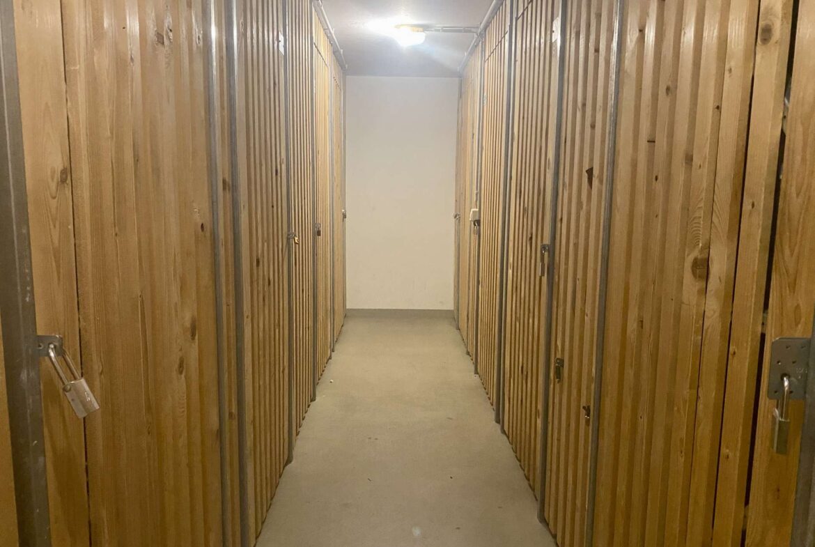 Narrow storage-unit hallway with wooden doors, metal frames, and padlocks along both sides, lit by a ceiling light.