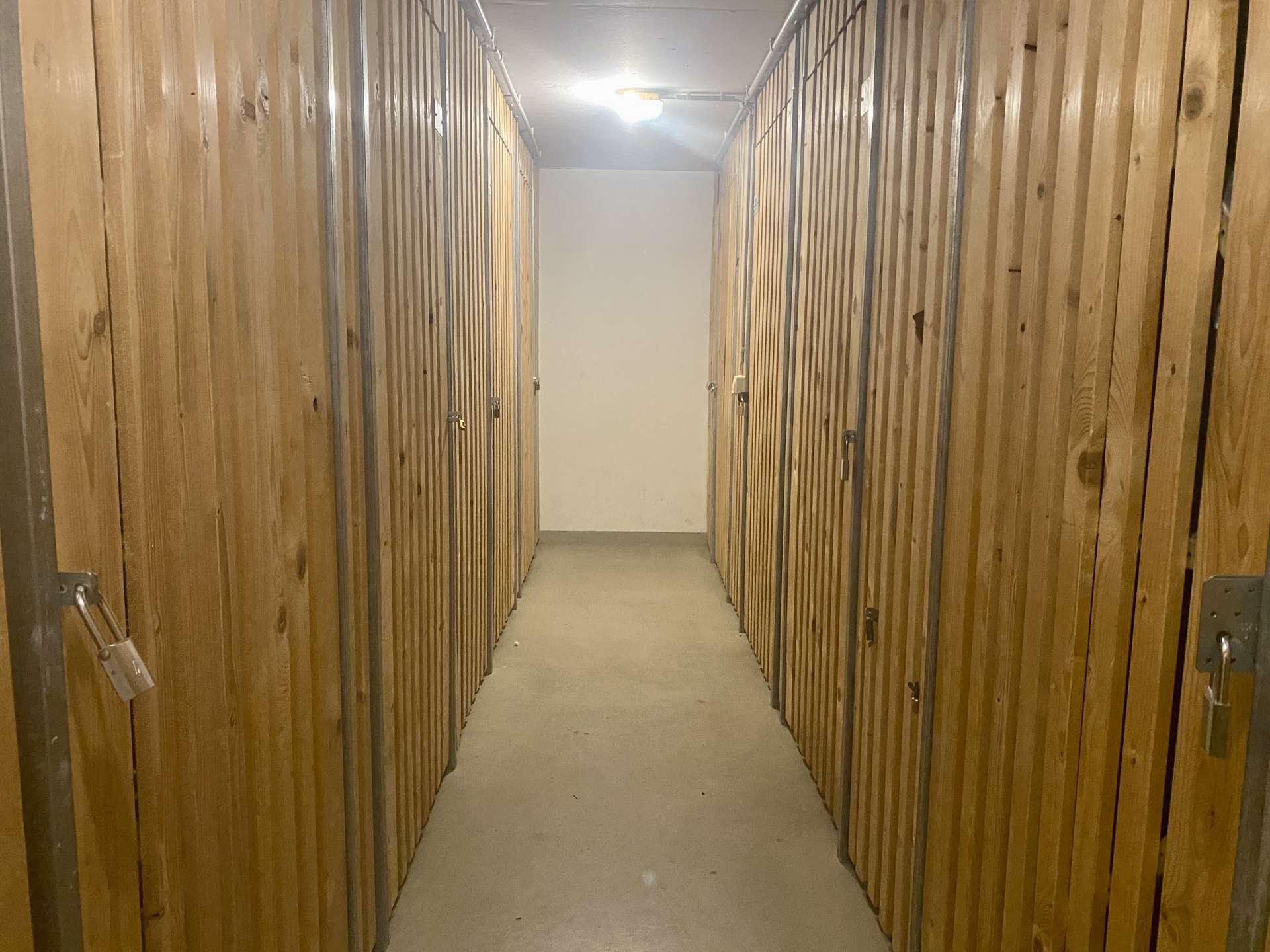 Narrow storage-unit hallway with wooden doors, metal frames, and padlocks along both sides, lit by a ceiling light.