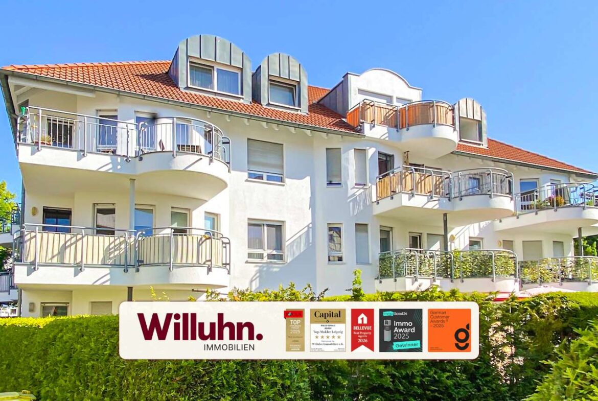 White multi‑story apartment building with curved balconies and a red-tiled roof; foreground shows a Willuhn Immobilien sign.