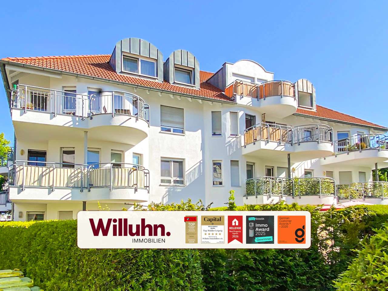White multi‑story apartment building with curved balconies and a red-tiled roof; foreground shows a Willuhn Immobilien sign.