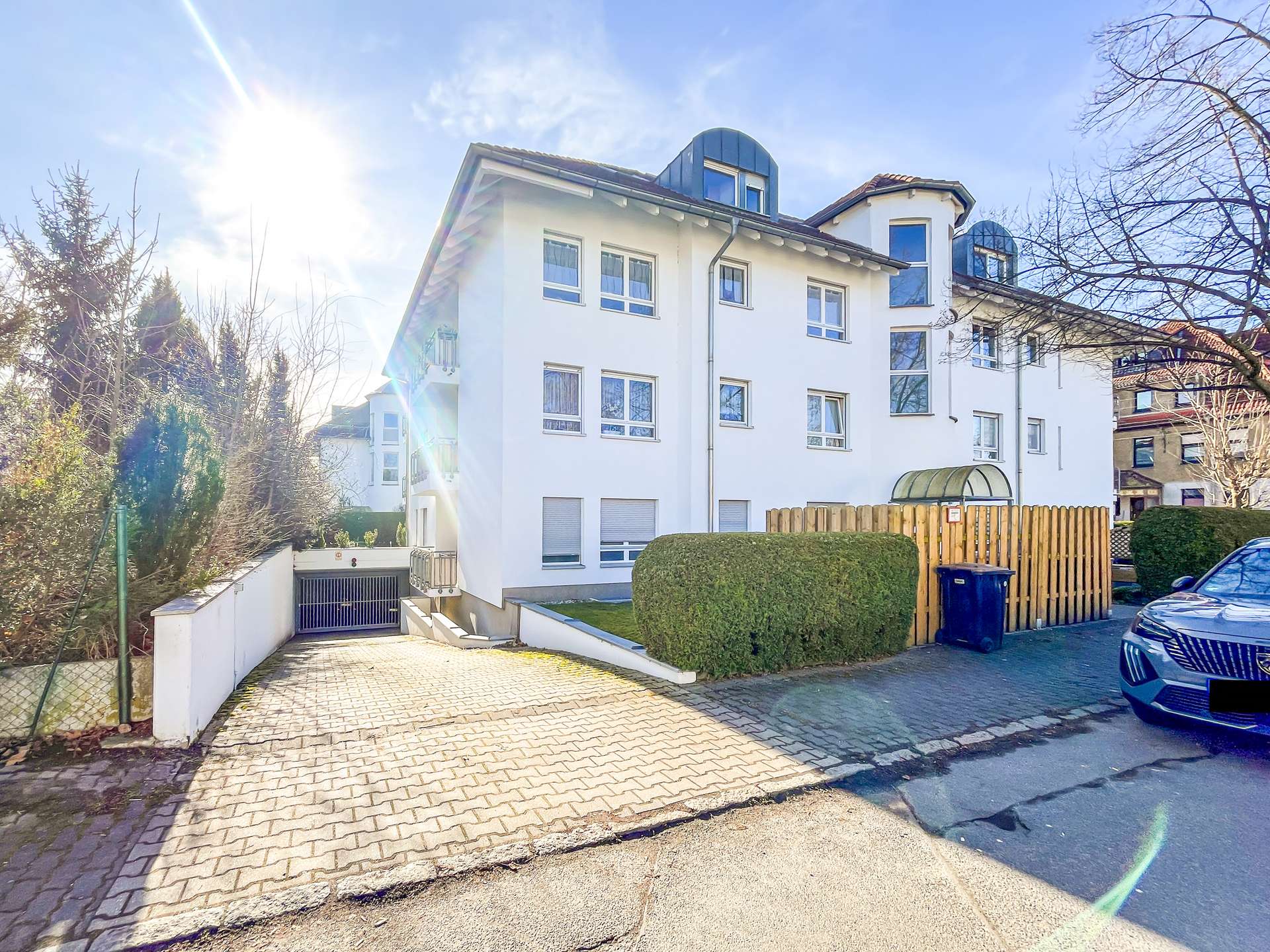 White multi-story residential building with blue-topped dormer windows, front courtyard, and a wooden fence on a sunny day. Full-page: White apartment building with gated driveway and parked car on the right, sun glare.