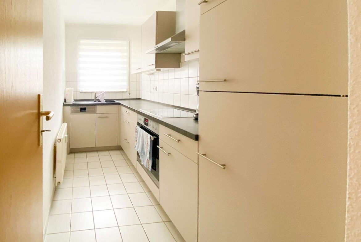 Narrow white kitchen with gray countertops, built-in oven and stove, sink under a window with blinds and tiled floor.