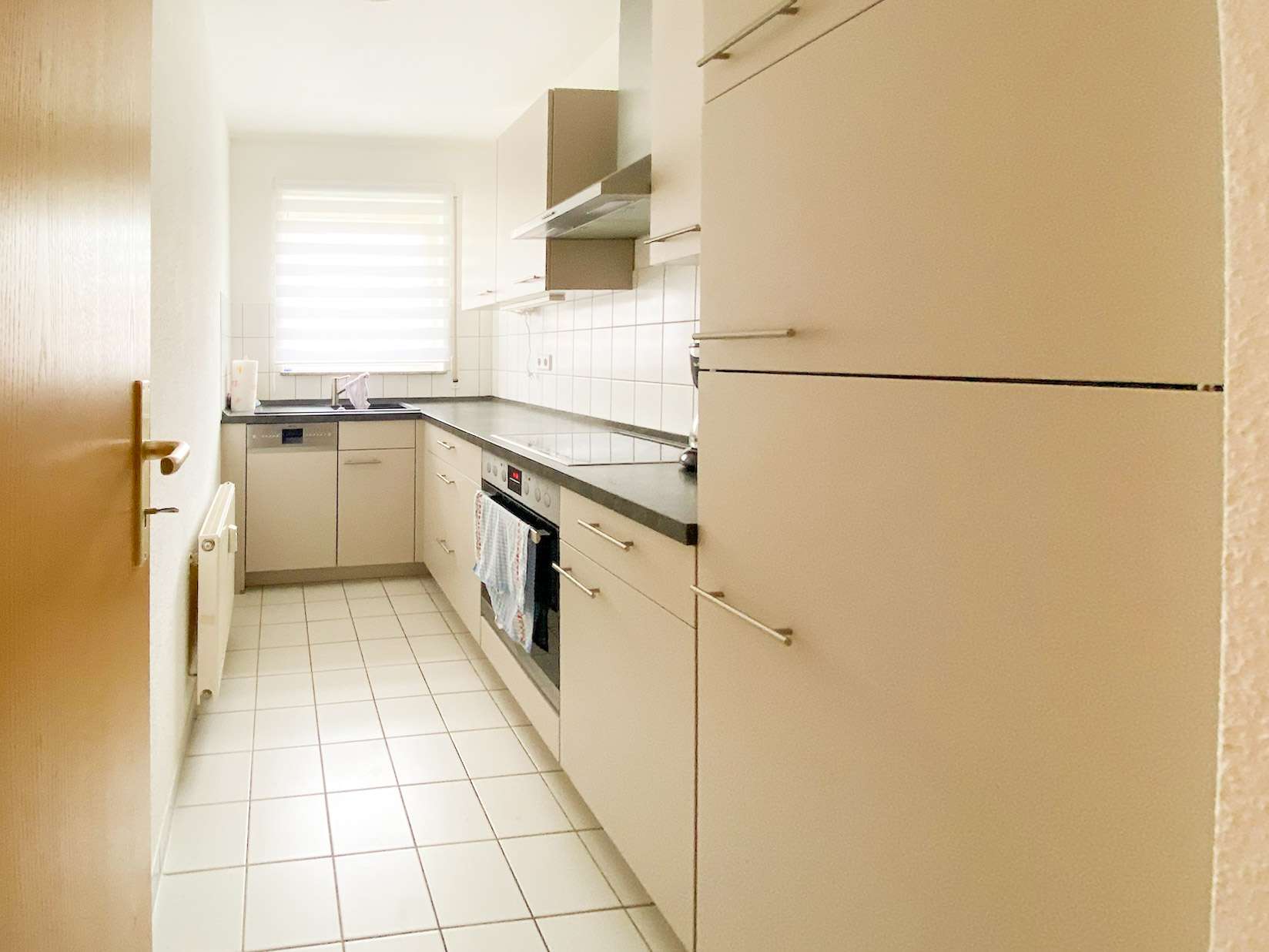 Narrow white kitchen with gray countertops, built-in oven and stove, sink under a window with blinds and tiled floor.