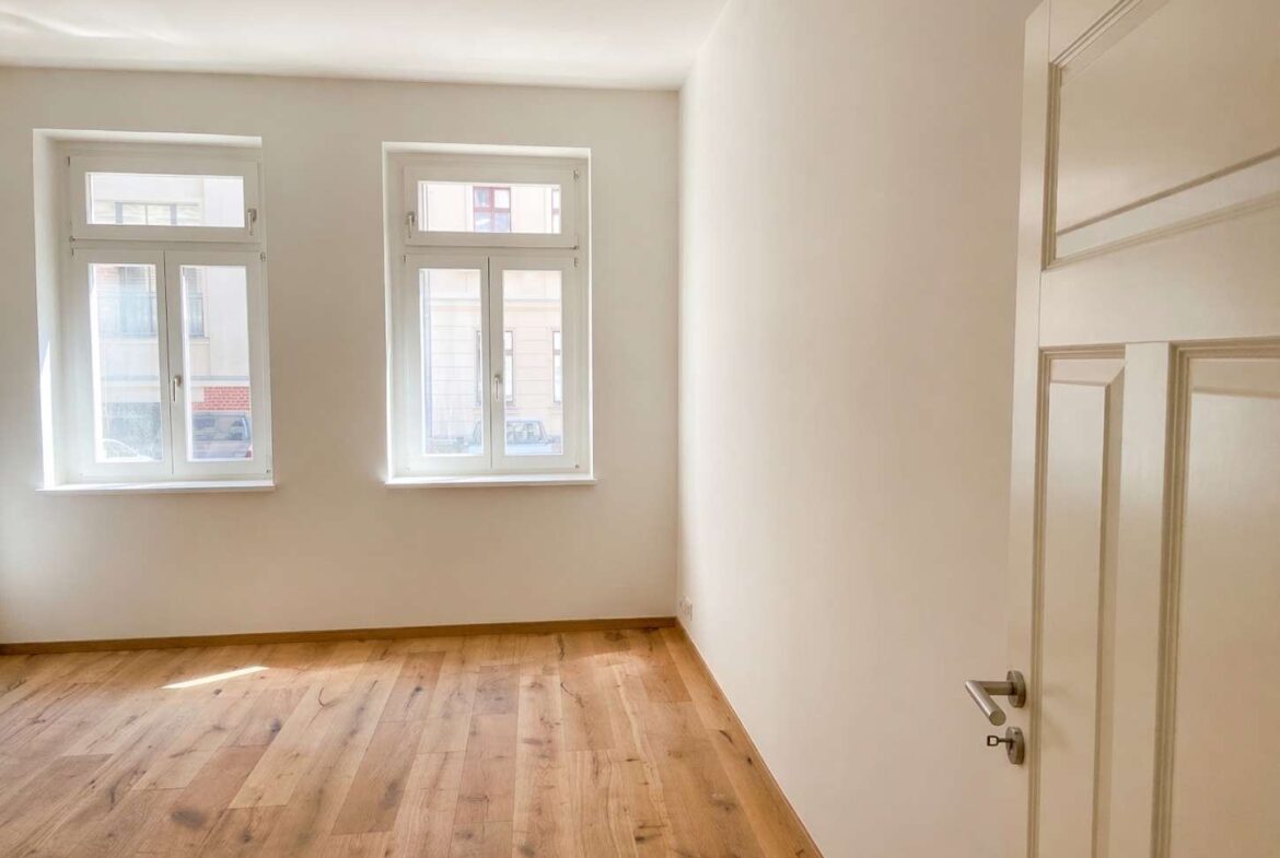 Empty room with light wooden floor, white walls, two tall windows letting in sunlight, and a white door on the right.