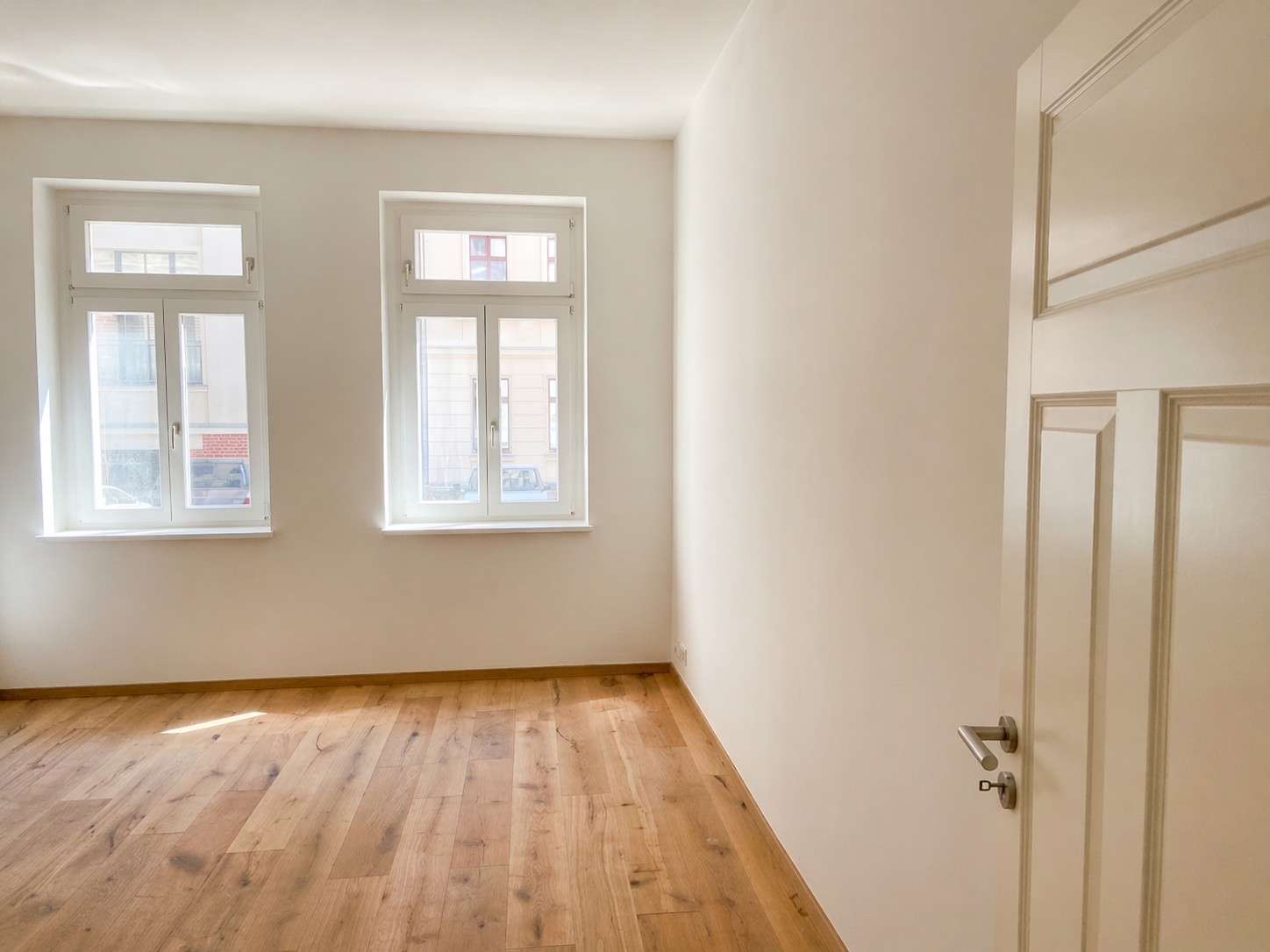Empty room with light wooden floor, white walls, two tall windows letting in sunlight, and a white door on the right.