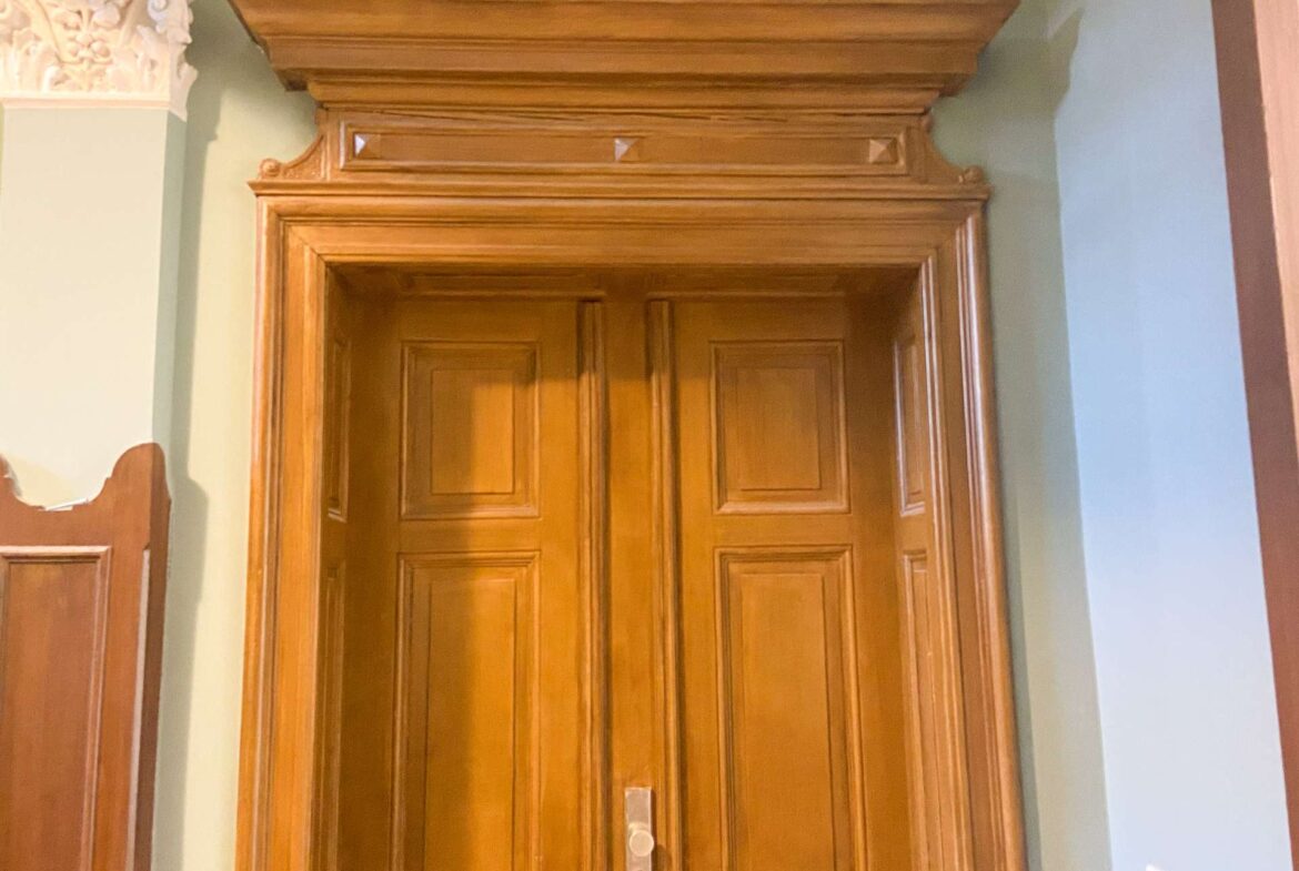 Tall wooden door with raised panels and an ornate frame, set in a green wall with damaged plaster visible on the left.