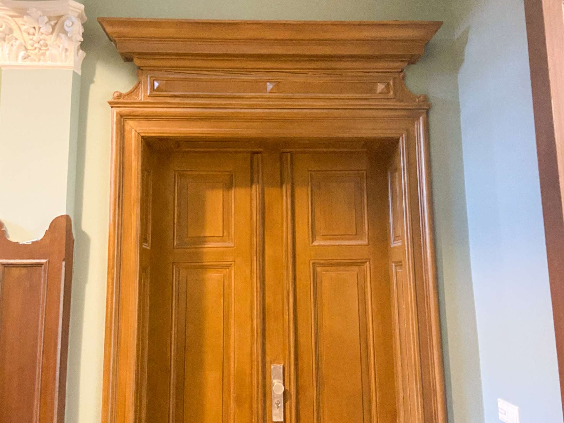 Tall wooden door with raised panels and an ornate frame, set in a green wall with damaged plaster visible on the left.