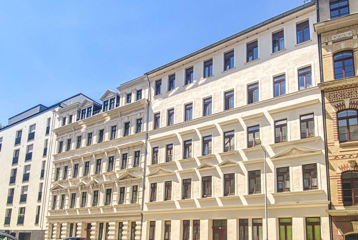 Row of light-colored apartment buildings with many windows along a sunny street, cars parked in front