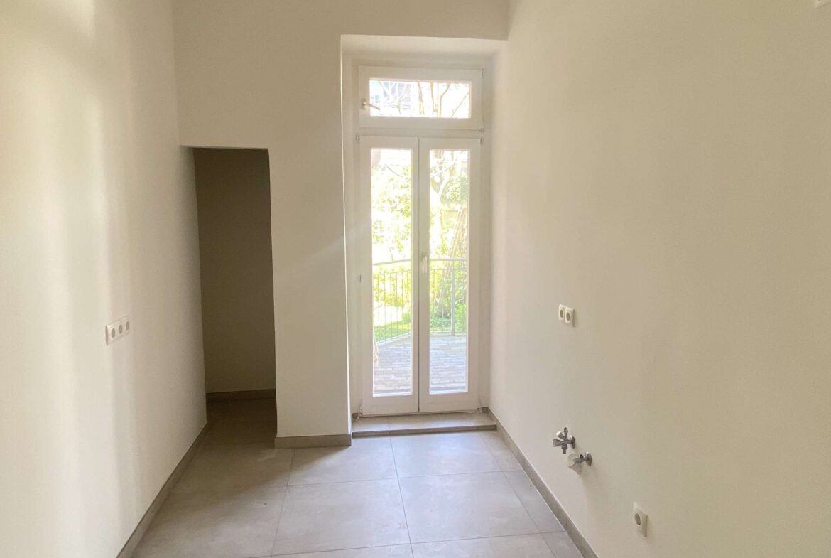 Narrow white utility room with a floor-to-ceiling windowed door and balcony outside, bright daylight in the space, plumbing visible on the right side.