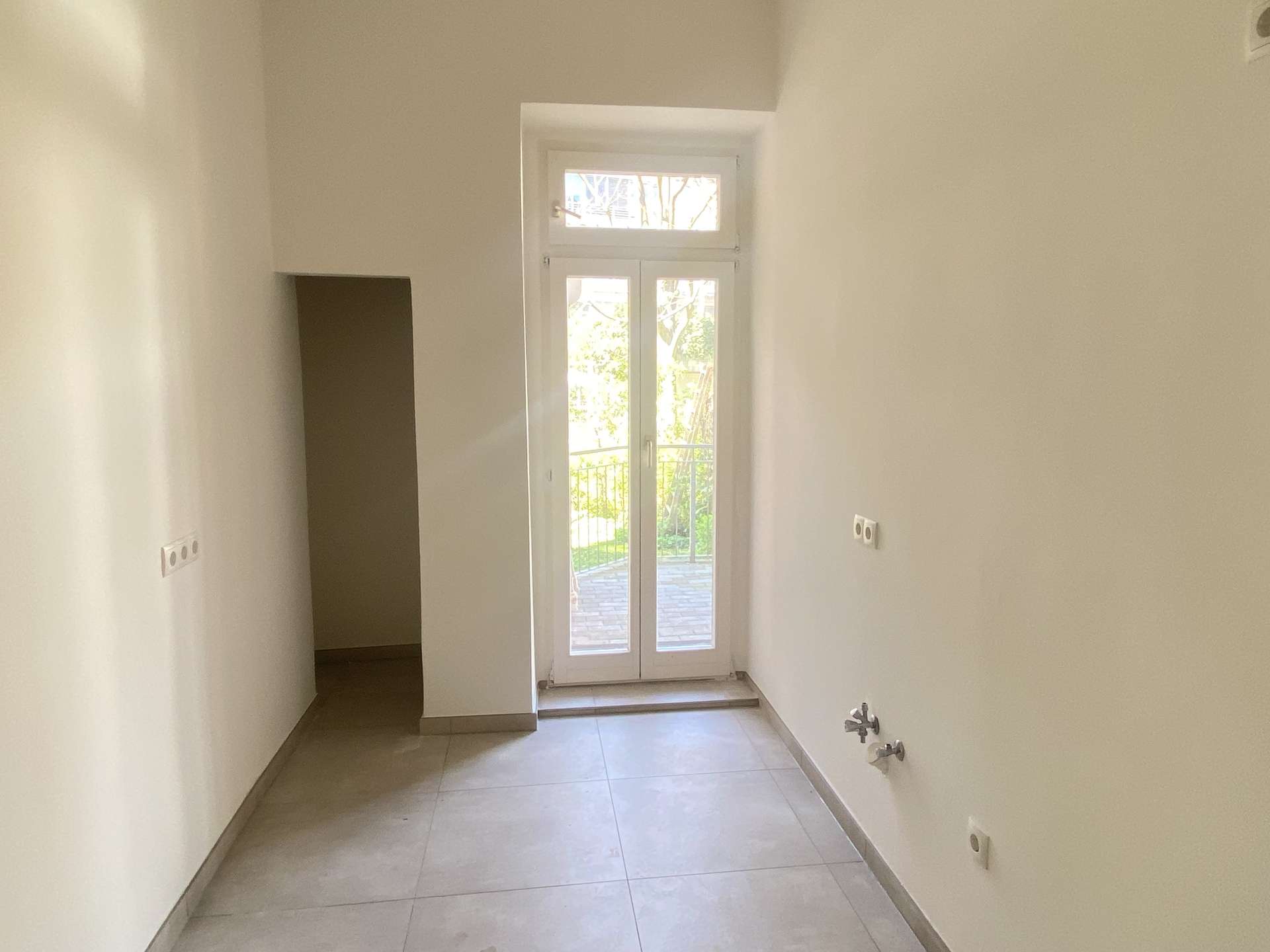 Narrow white utility room with a floor-to-ceiling windowed door and balcony outside, bright daylight in the space, plumbing visible on the right side.