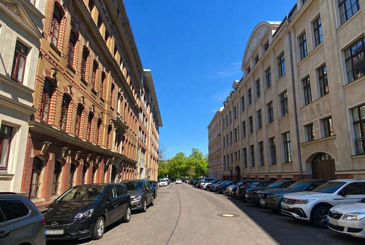 Street between brick left building and pale right building, with parked cars along both curbs and a clear blue sky above.