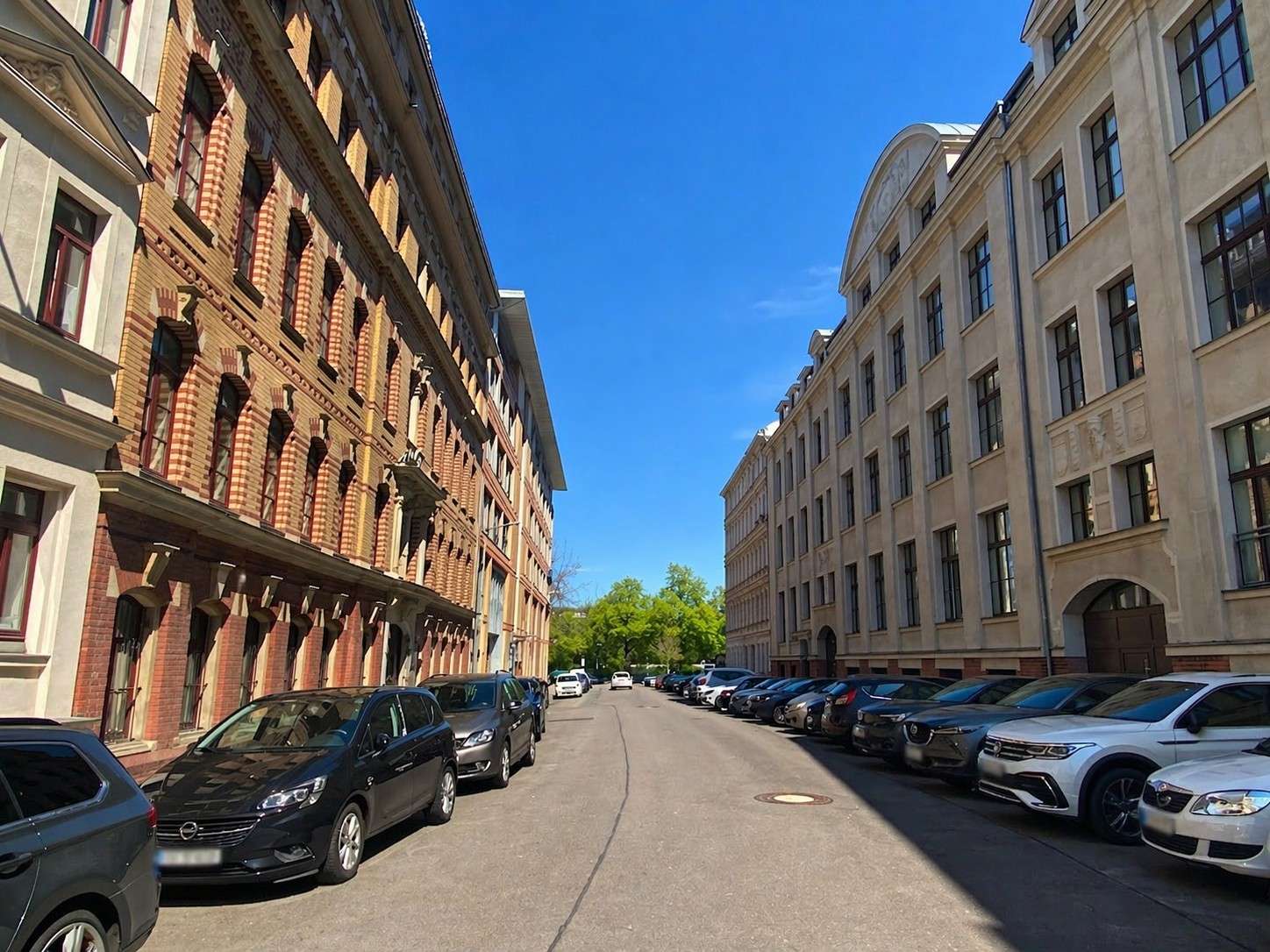 Street between brick left building and pale right building, with parked cars along both curbs and a clear blue sky above.