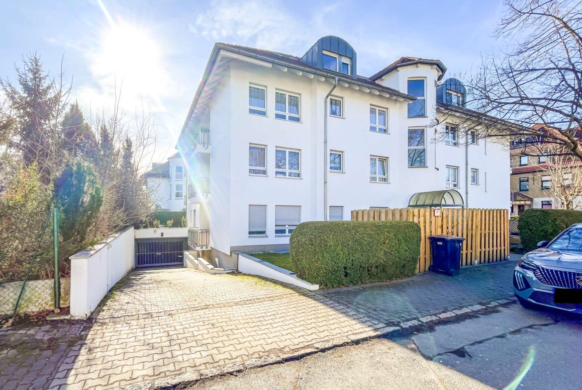 White multi-story apartment building with curved blue dormer windows, facing a cobblestone driveway on a sunny day, parked car on the right and a wooden fence in front.