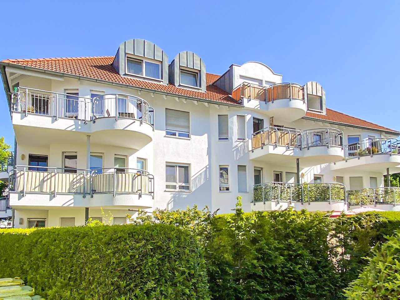 White modern apartment building with curved balconies, red-tiled roof, and a blue sky above.