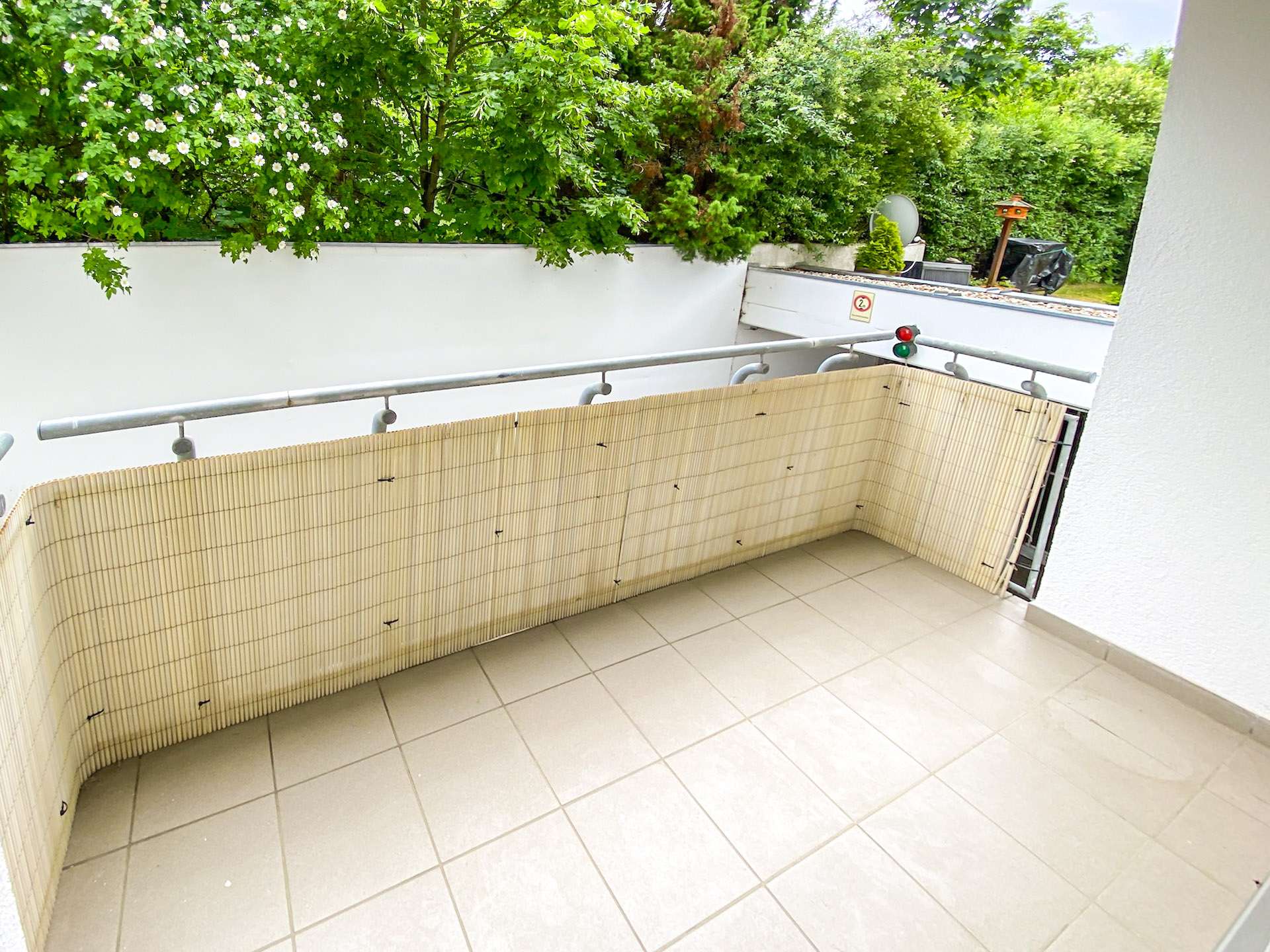 Empty tiled balcony with a beige bamboo privacy screen along the railing and green shrubs beyond.