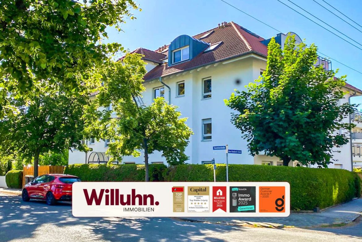 White multi-story residential building with a red-tiled roof, trees in front, and a large Willuhn Immobilien banner with award badges in the foreground.