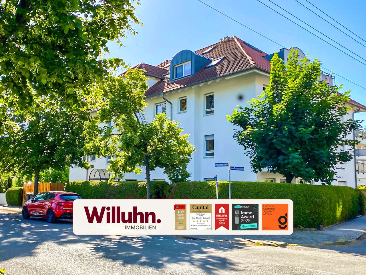 White multi-story residential building with a red-tiled roof, trees in front, and a large Willuhn Immobilien banner with award badges in the foreground.