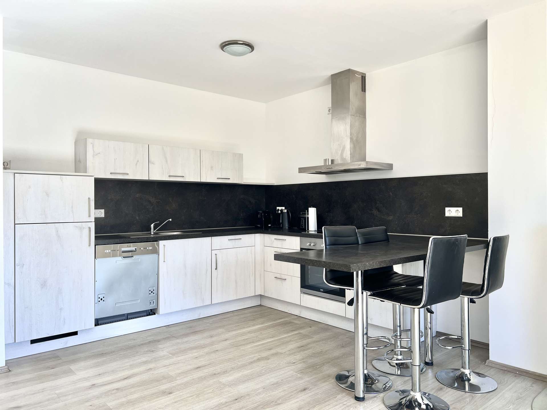 Modern L-shaped kitchen with white cabinets, black backsplash, stainless appliances, and a tall island with chrome bar stools.