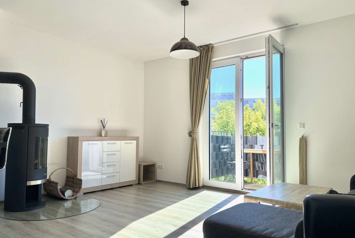 Living room with a black wood stove on the left, light wood cabinet, and a glass door opening to a balcony bathed in sunlight.