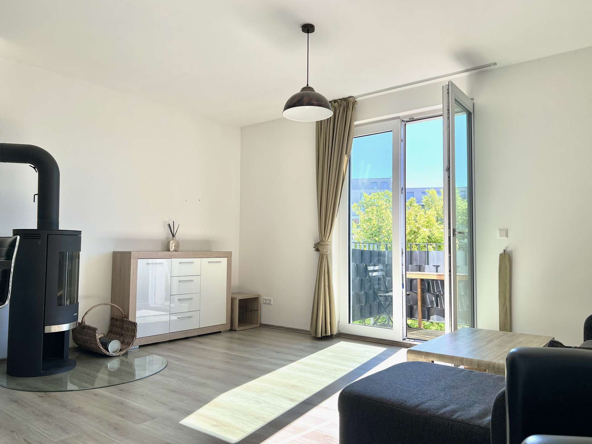 Living room with a black wood stove on the left, light wood cabinet, and a glass door opening to a balcony bathed in sunlight.