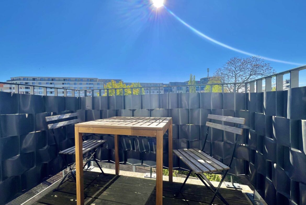 Rooftop terrace with a wooden table and benches, surrounded by dark metal privacy panels against a bright blue sky with sun glare.