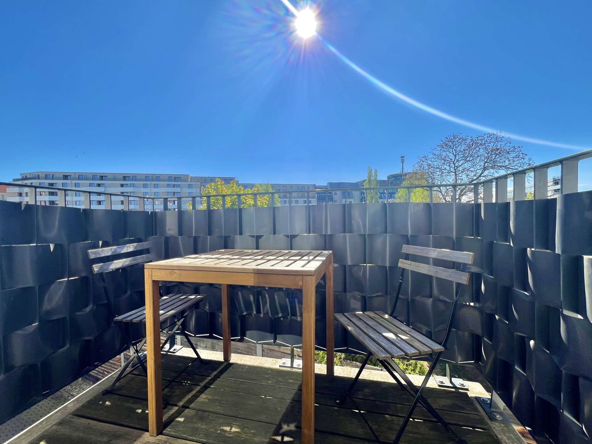 Rooftop terrace with a wooden table and benches, surrounded by dark metal privacy panels against a bright blue sky with sun glare.