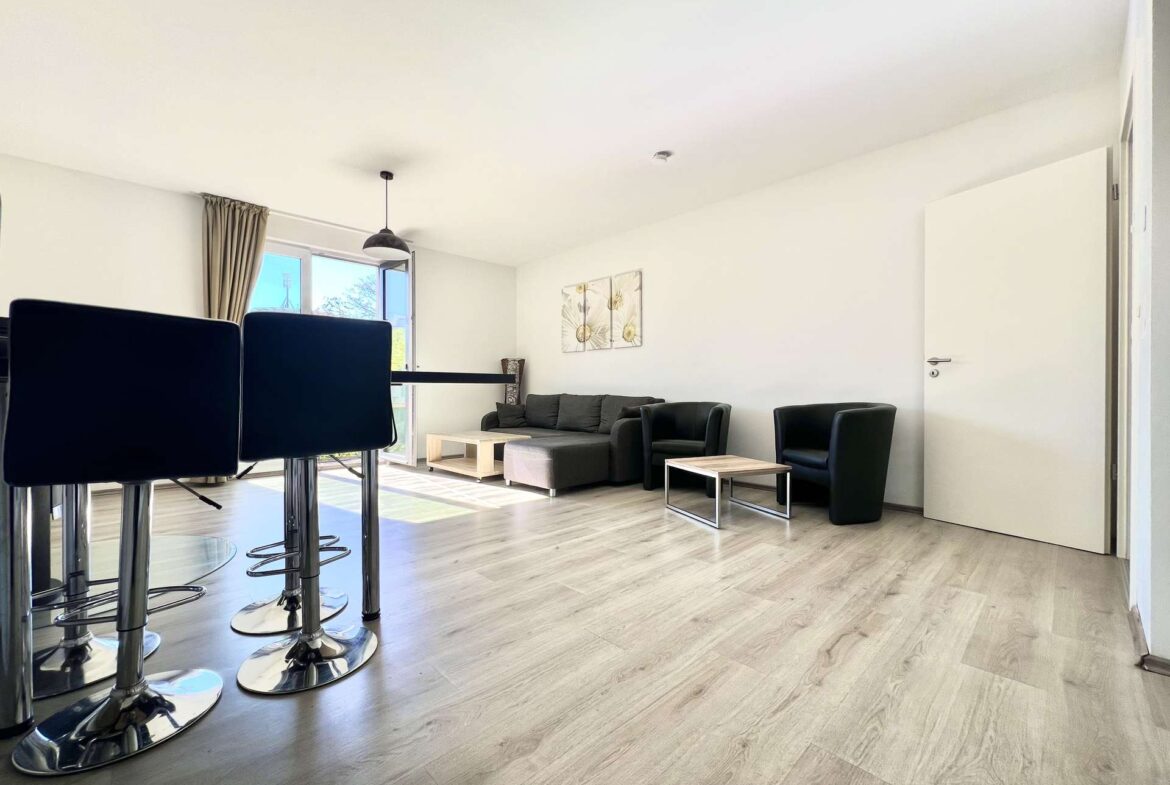 Open-plan living room with black bar stools in the foreground, light wood floor, white walls, gray sofa, and glass doors letting in sunlight.