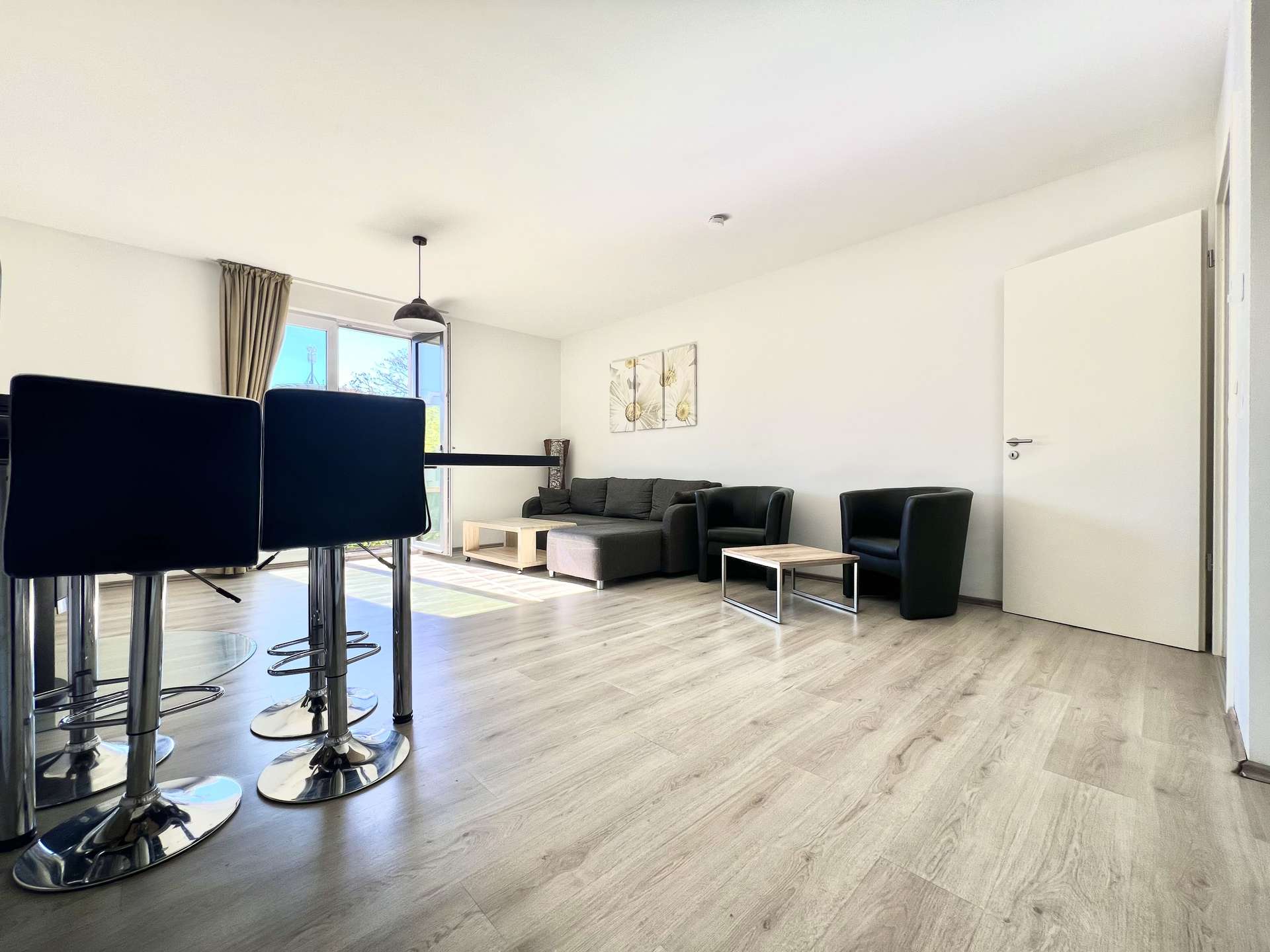 Open-plan living room with black bar stools in the foreground, light wood floor, white walls, gray sofa, and glass doors letting in sunlight.