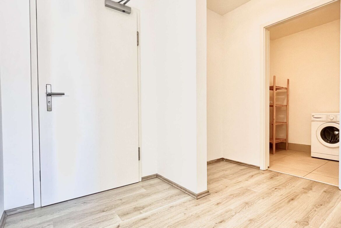 White hallway with closed white doors on the left and an open doorway to a laundry room on the right, showing a washing machine and a wooden shelf unit.